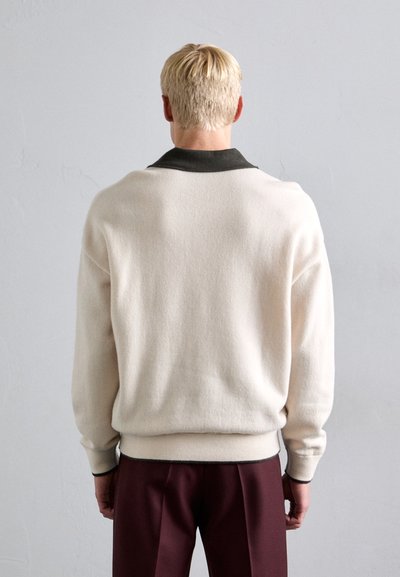 Persoon met kort blond haar, gekleed in een crèmekleurige trui met een zwarte kraag en bordeauxrode broek, staand voor een lichtgrijze muur.