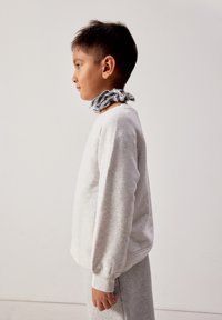 Garçon portant un sweat-shirt gris et un pantalon, debout de profil, avec une écharpe à carreaux nouée autour du cou contre un fond uni.
