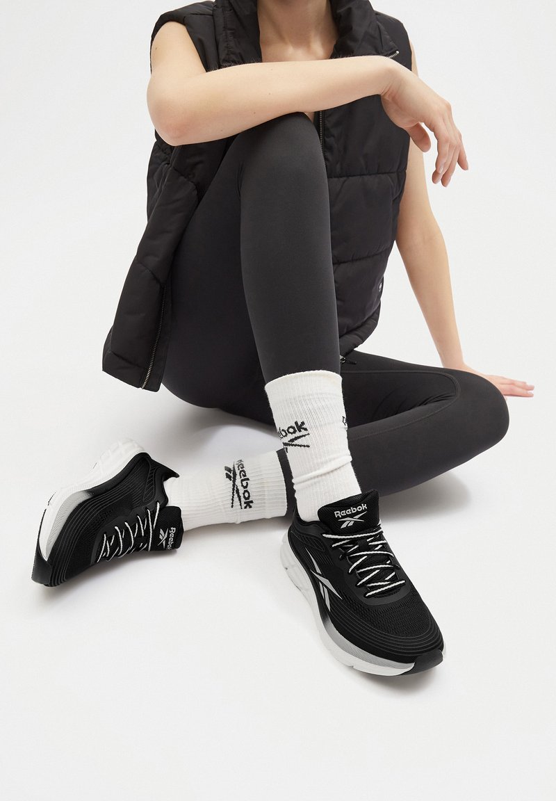 Fekete sportcipők, textúrált hálós felsőrésszel, fehér talppal és fűzőkkel. Fehér zoknikkal viselve, amelyeken a Reebok logó látható, valamint fekete leggings-szel.