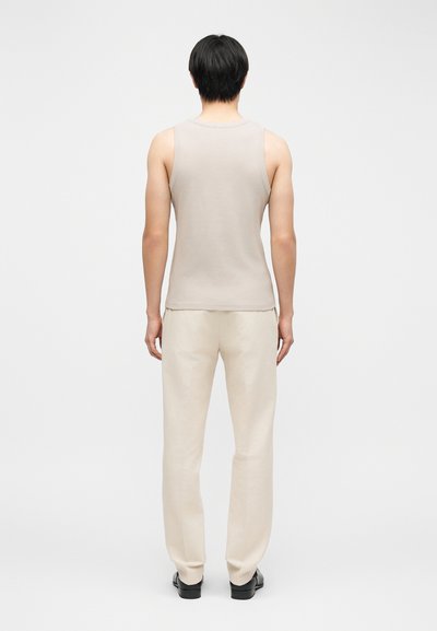 Άνδρας που στέκεται γυρισμένος από την πλάτη, φορώντας μπεζ αμάνικο τοπ, παντελόνι κρεμ και μαύρα παπούτσια, σε απλό φόντο.