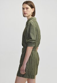 Tuta verde oliva con un tessuto testurizzato, maniche corte, colletto con bottoni e vita elasticizzata, caratterizzata da righe verticali.