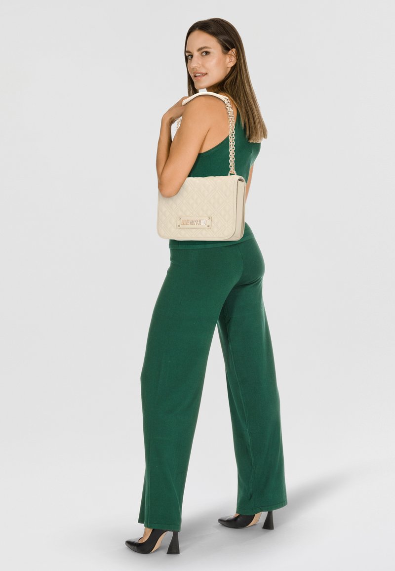 Tuta verde con top aderente e gambe ampie, abbinato a una borsa a tracolla beige trapuntata con catena e dettagli metallici. Tacchi alti neri.