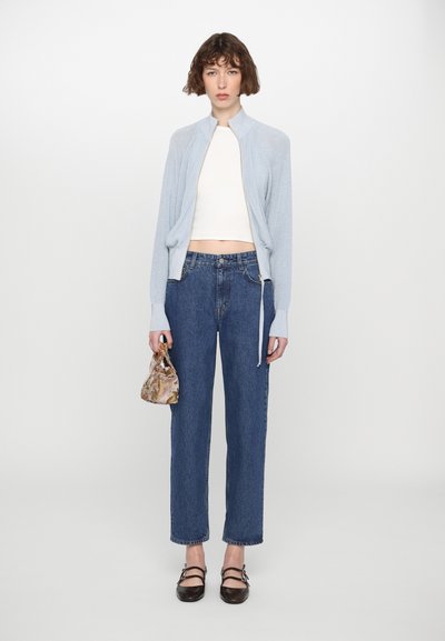Cárdigan azul claro con cremallera, top blanco corto, vaqueros azul de cintura alta, bolso pequeño con estampado y zapatos planos negros con doble tirante.