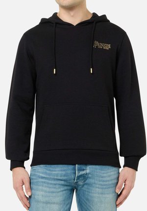 Mann trägt schwarzen Hoodie mit goldenem Logo auf der Brust und blaue Jeans vor weißem Hintergrund.