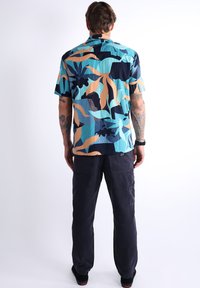 Chemise à manches courtes avec des motifs de feuilles abstraits en bleu, noir et beige. Le tissu a une surface texturée, associée à un pantalon sombre.