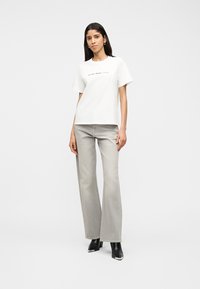 Γυναίκα που στέκεται, φορά λευκό T-shirt με κείμενο, ανοιχτό γκρι τζιν με φαρδύ πόδι και μαύρα μπότες με τακούνι, μπροστά σε λευκό φόντο.