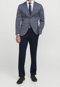 Uomo che indossa un blazer grigio a quadri, camicia bianca, cravatta blu navy, pantaloni blu scuro, cintura marrone e scarpe nere, in piedi con una mano in tasca.