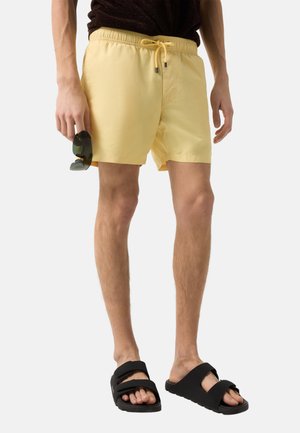 Mand iført lysegule badeshorts, holder solbriller og sorte slidesandaler, stående mod en ensfarvet lys baggrund.