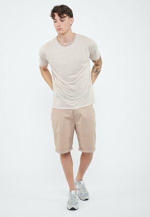 Giovane uomo con capelli castani corti che indossa una maglietta beige a righe, pantaloncini beige risvoltati, sneakers bianche, in piedi con le mani dietro la schiena.