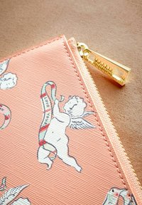 Puzdro v broskyňovej farbe so zúbkovou textúrou, s ilustráciami cherubínov a akcentmi v tvare cukrovej paličky; obsahuje detail zlatého zipsu.
