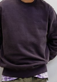 Ciemnofioletowa bluza wykonana z mieszanki bawełny, z okrągłym dekoltem, ściągaczami na rękawach oraz wytłoczonym logo na piersi. Noszona na warstwę z bluzką tie-dye.