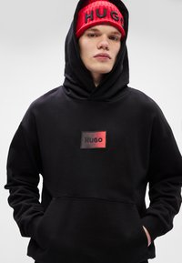 Μαύρο φούτερ με κουκούλα, που παρουσιάζει ένα κεντρικό ορθογώνιο λογότυπο με την ένδειξη "HUGO" σε έντονη κόκκινη και μαύρη γραφή. Μαλακό ύφασμα, καθημερινή εφαρμογή.