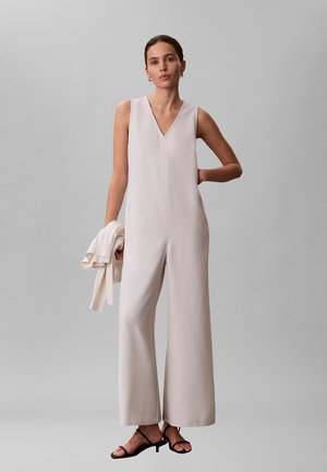 Mujer con mono beige sin mangas y sandalias negras de tacón, sosteniendo un suéter blanco doblado en una mano sobre un fondo gris liso.