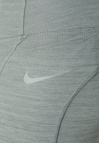 Leggings sportivi Nike in tessuto grigio chiaro con una superficie liscia. Presentano un piccolo logo riflettente e cuciture rinforzate per la durata.