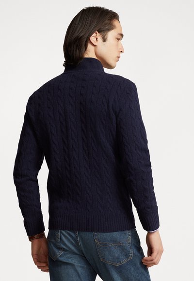 Mørkeblå kabelstrikket trøje med høj krave og ribbede håndled, der viser et tekstureret mønster. Båret med blå jeans, set fra ryggen.