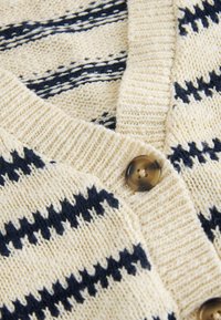 Cardigan tricoté crème avec des rayures zigzag bleu marine et des boutons marron en écaille, montrant le col côtelé et un tissu texturé en gros plan.