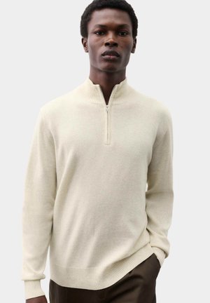 Man die staat met één hand in zijn zak, draagt een crèmekleurige half-zip trui en donkere broek, kijkt naar voren tegen een effen achtergrond.