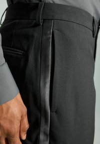 Pantalons noirs à la coupe ajustée en tissu lisse, dotés d'une bande satinée sur le côté, de détails de plis et d'une poche discrète. Accompagnés d'une chemise grise.