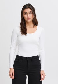 Hosszú ujjú fehér felső lekerekített nyakkivágással, fekete nadrággal párosítva. Az anyag sima és testhezálló hatású.