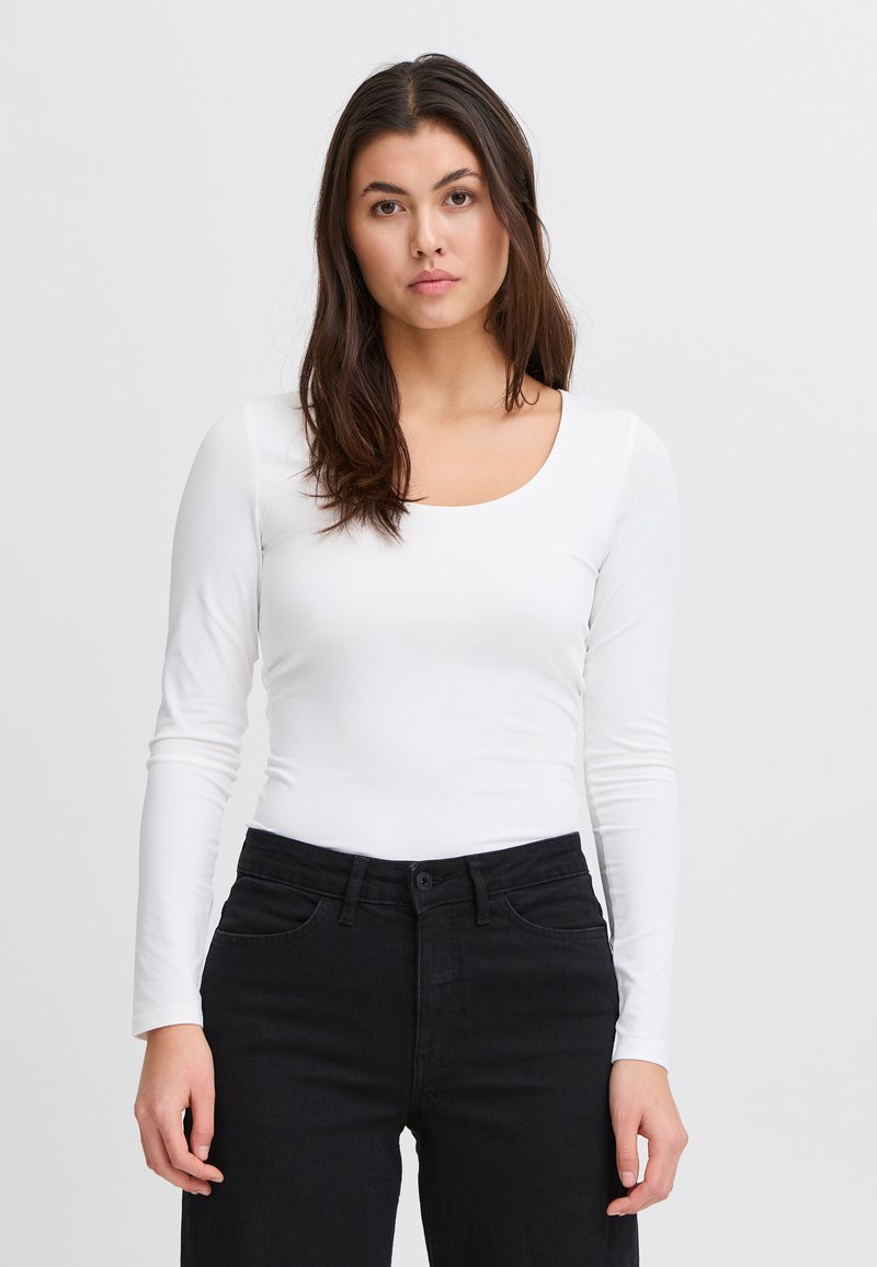 Hosszú ujjú fehér felső lekerekített nyakkivágással, fekete nadrággal párosítva. Az anyag sima és testhezálló hatású.