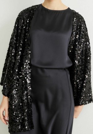 Femme portant une robe en satin noire associée à une veste noire ouverte à l'avant ornée de sequins, présentée du cou jusqu'à mi-cuisse sur un fond uni.