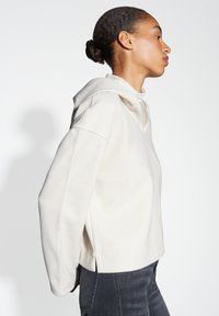 Felpa con cappuccio in morbido tessuto color crema chiaro. Design ampio con spalle scese, fessure laterali e dettagli a coste sui polsini.