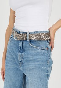 Ruskea kudottu vyö, jossa on teksturoitu kuvio ja hopeinen metallisolki, käytetty vaaleansinisten, rennosti istuvien farkkujen kanssa.