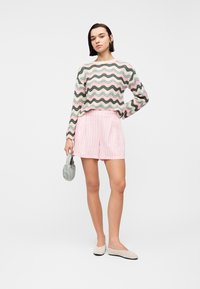 Νέα γυναίκα που φοράει πολύχρωμο πουλόβερ με μακριά μανίκια, ροζ ριγέ σορτς, μπεζ μπαλαρίνες και κρατά ένα μικρό ανοιχτόχρωμο τσαντάκι.