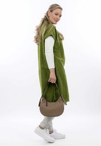 Abito smanicato oversize in verde con texture, camicia a maniche lunghe bianca sotto, borsa a tracolla beige, pantaloni grigi chiari, sneaker bianche.