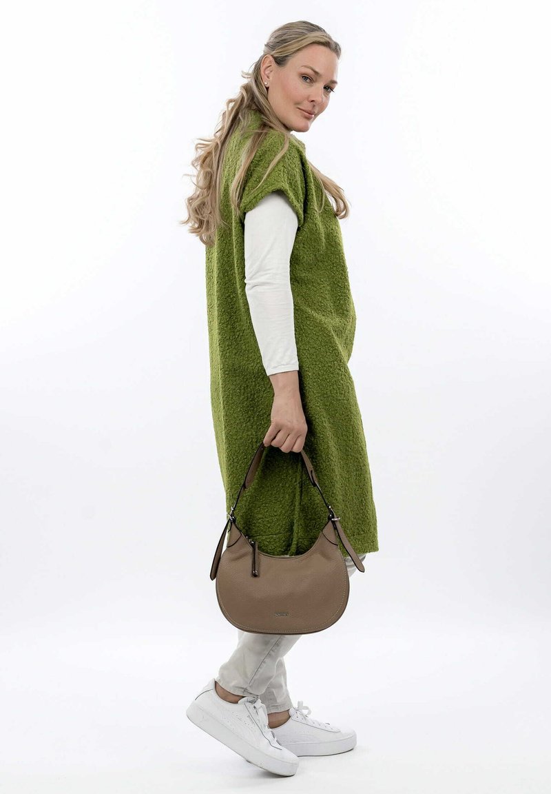 Abito smanicato oversize in verde con texture, camicia a maniche lunghe bianca sotto, borsa a tracolla beige, pantaloni grigi chiari, sneaker bianche.