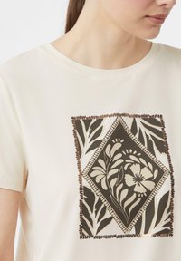 Jasno-beżowa koszulka z centralnym wzorem diamentu florystycznego w kolorze brązowym, wykończona dekoracyjnym haftem i otoczona liściastymi wzorami.