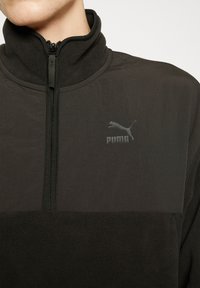 Svart zip-up fleece-pullover med hög krage, med en slät övre panel och en texturerad nedre panel. Puma-logotyp i svart.