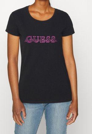 Kobieta ubrana w czarny krótki T-shirt z różowym napisem "GUESS" i jasnoniebieskie dżinsy na jednolitym białym tle.