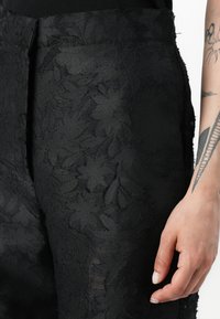 Black floral textured pants with a smooth finish, featuring an intricate leaf pattern and a fitted silhouette. Tapered leg design.