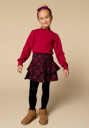 Rode langmouwtshirt met ruches bij de hals, gecombineerd met een zwarte gelaagde rok met roze hartpatronen, zwarte leggings en bruine laarzen.