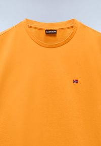 Tricou din bumbac portocalie cu guler rotund. Prezintă o mică broderie în formă de steag pe piept. Textură netedă, design minimalist și mâneci scurte.