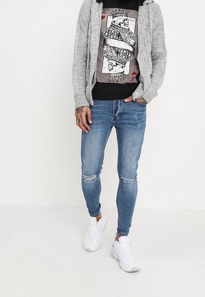 Grau gestrickte Strickjacke über einem schwarzen Grafik-T-Shirt. Verwaschene blaue Skinny-Jeans mit abgenutzten Knien. Weiße Turnschuhe runden das Outfit ab.