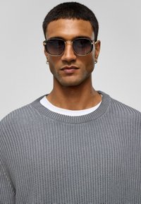 Zonnebril met ronde donkere lenzen, gouden metalen montuur. Model draagt een grijze gebreide trui over een witte shirt. Glad huid, kort haar.