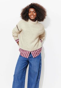 Pull en maille beige avec col côtelé et détails latéraux, porté par-dessus une chemise à rayures bordeaux boutonnée. Associé à un pantalon large en denim bleu.