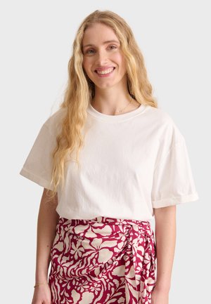 T-shirt bianco in cotone a maniche corte e vestibilità rilassata, indossato con una gonna bordeaux a fantasia con disegni floreali beige.
