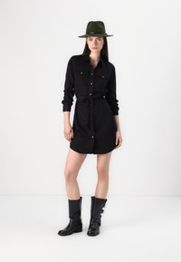 Rochie neagră cu curea la talie, cu nasturi în față și buzunare pe piept. Asortată cu cizme negre până la genunchi și o pălărie verde cu margine lată.
