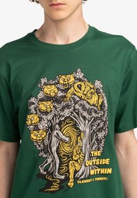 Camiseta verde de algodón con un diseño de naturaleza caricaturesco que incluye osos, árboles y un patrón en espiral. El texto impreso dice "EL EXTERIOR DENTRO".