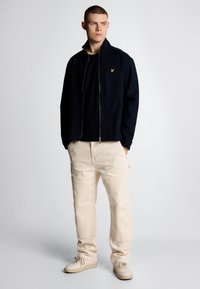 Giacca navy con collo alto e logo giallo, abbinata a pantaloni beige e sneakers beige, creando un outfit casual.