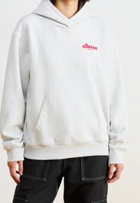 Persona che indossa una felpa grigia chiara delle ellesse con tasca frontale e pantaloni neri con cuciture bianche, in piedi contro uno sfondo neutro.