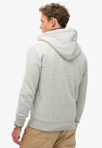 Man met krullend haar, gekleed in een lichtgrijze hoodie en beige broek, van achter bekeken met zijn hoofd naar links gedraaid.