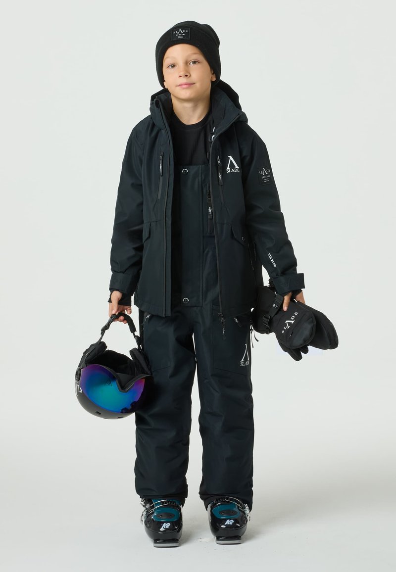 Zwart skioutfit bestaande uit een jas en een overall, textiel met structuur, voorzien van logo's en ritsen. Accessoires: helm met een iriserende vizier, handschoenen.