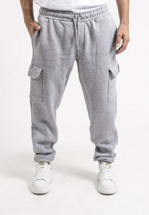 Homme portant un pantalon de survêtement cargo gris clair avec une ceinture élastique et des baskets blanches, debout avec les mains près des poches.