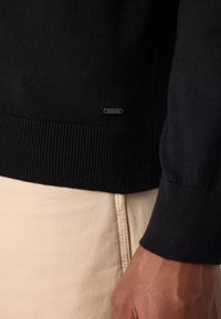 Pull noir en tricot avec ourlet côtelé, arborant une petite étiquette logo. Porté sur un tissu beige clair, mettant en valeur la texture et la coupe.