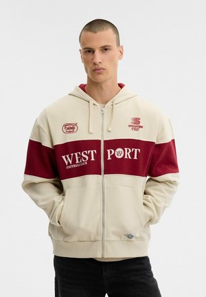 Un modelo masculino está de pie sobre un fondo blanco, con una sudadera con capucha y cremallera de color crema, paneles granates y el texto «West Port Football Club».