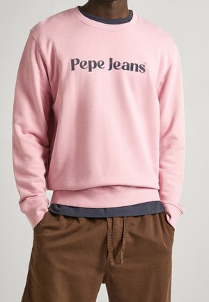 Homme portant un sweat-shirt rose Pepe Jeans sur une chemise sombre et un pantalon marron, mains dans les poches, sur fond uni.